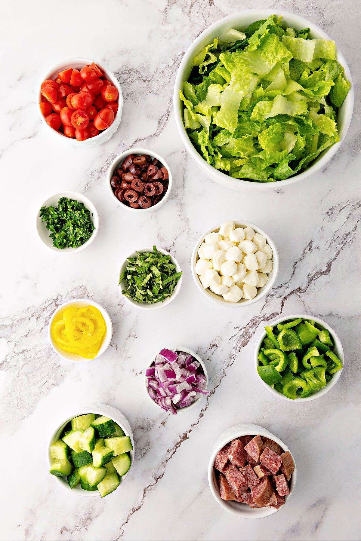 Bowl of lettuce, mozzarella pearls, cherry tomatoes, cucumbers, banana peppers, herbs, olives, bell peppers, and chopped meat.