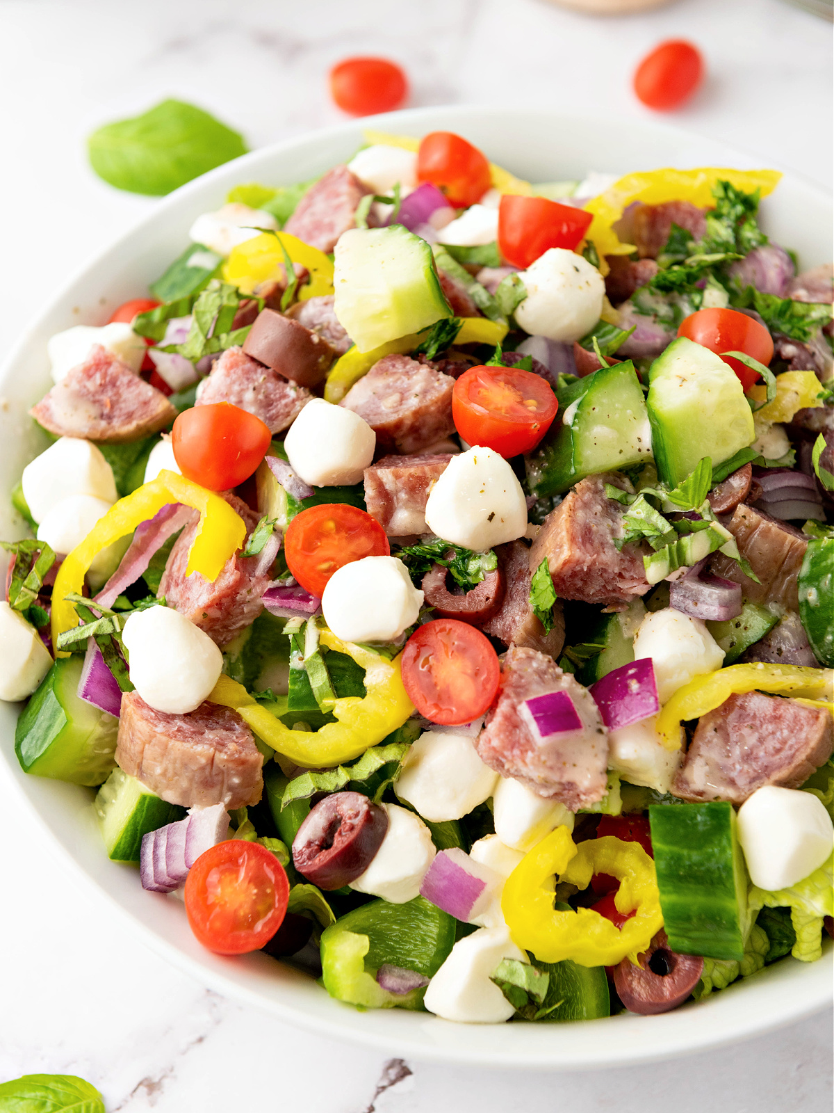 Bowl of chopped salad with mozzarella pearls, cherry tomatoes, cucumbers, banana peppers, and chopped meat.