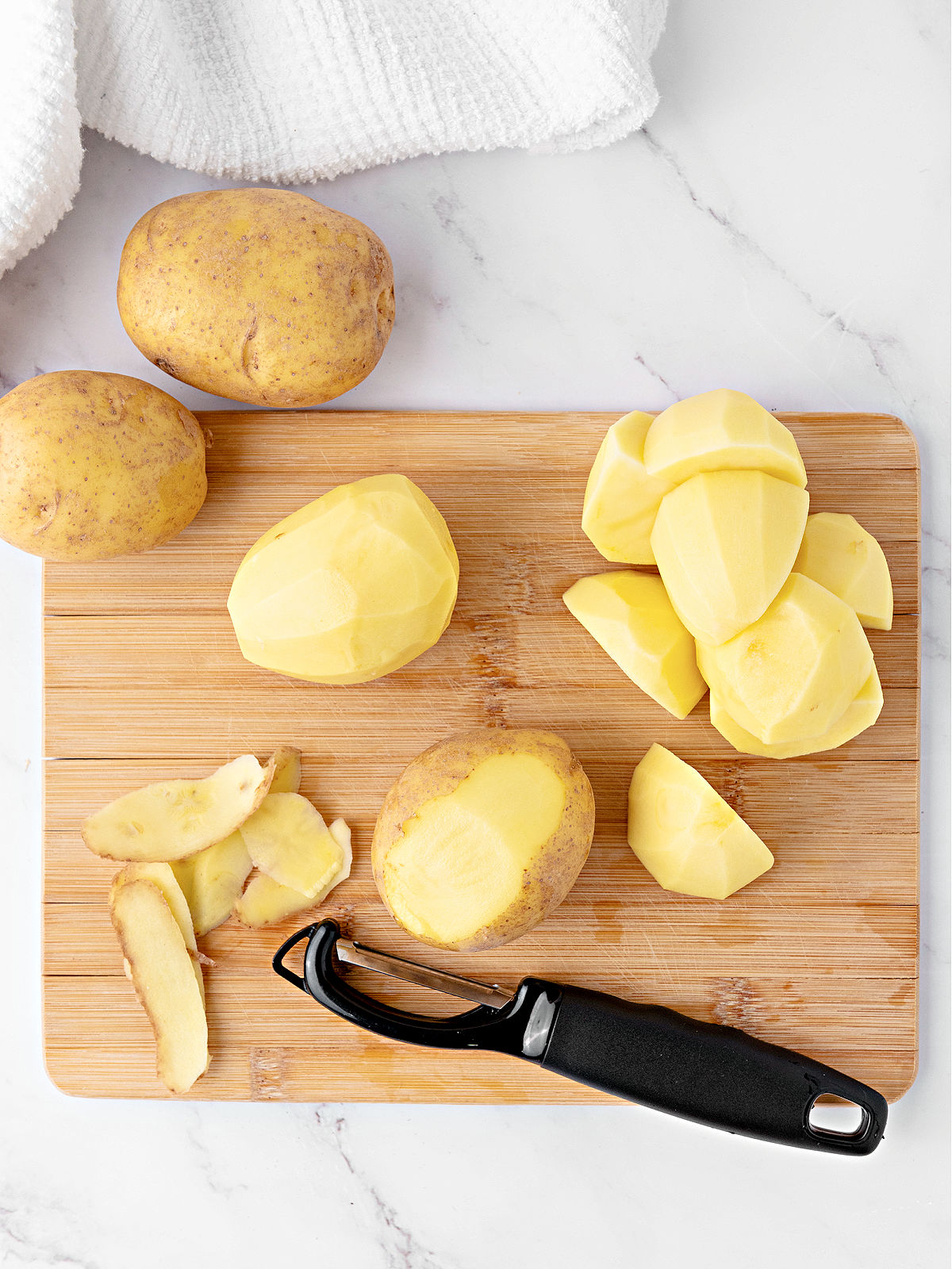 Peeled potatoes and a peeler on a cutting board.