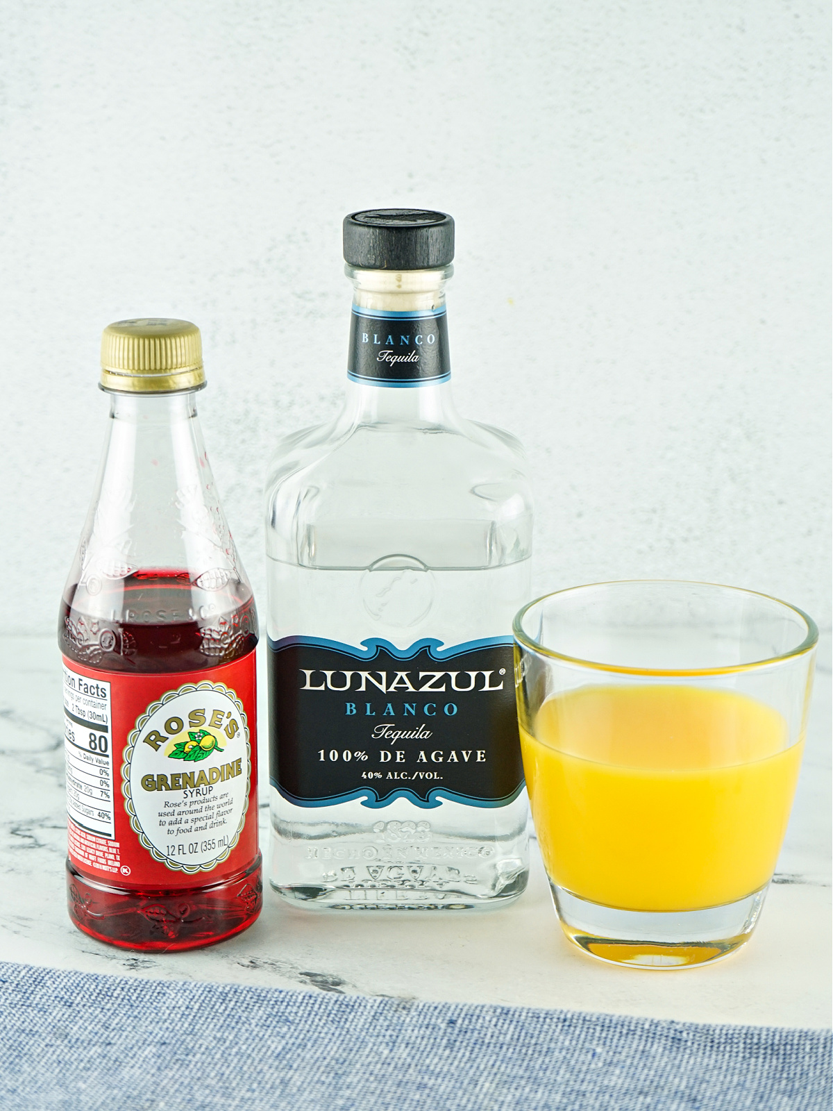 A glass of orange juice and bottles of grenadine syrup and white tequila.