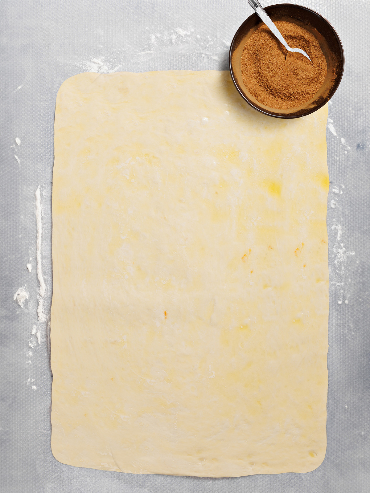 Dough rolled into a rectangle, covered with butter, and a small bowl of cinnamon sugar.