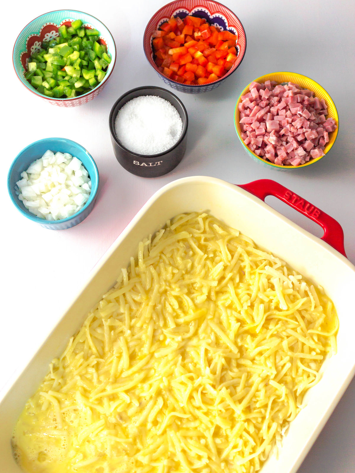 Hash browns and eggs in a baking dish and ham, peppers, onions, and salt in small bowls.