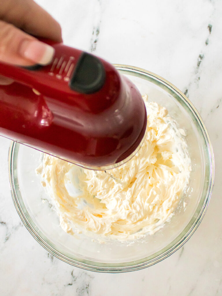 Red handheld mixer beating cream cheese mixture in a glass bowl.